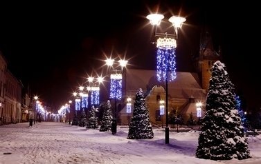 Zdjęcie przedstawia czarnkowski deptak w zimowej scenerii, długa ulica, biegnie r&oacute;wnolegle do zabudowań mieszkalnych z lewej strony i oświetlenia ulicznego z prawej, pomiędzy lampami ulicznymi dekoracje bożonarodzeniowe