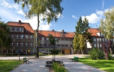 Dyży murowany, trzykondygnacyjny budynek szkoły, pokryty czerwona dach&oacute;wką, na pierwszym planie chodnik i pojedyncze drzewo