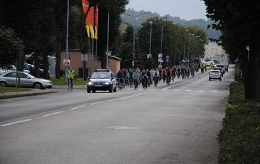 Rajd rowerowy ulicami Czarnkowa. Jedną z gł&oacute;wnych ulic miasta jadą rowerzyści, przed nimi radiow&oacute;z policyjny.