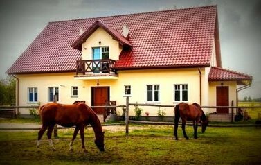 Zdjęcie przedstawia Chatę MIłkowską. Duży dom jednorodzinny z czerwonym dachem. Przed domem pasą się dwa gniade konie.