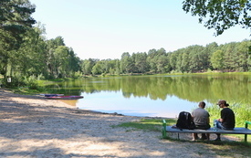 plaża przy jeziorze. Nad jeziorem turyści siedzą na ławeczce.